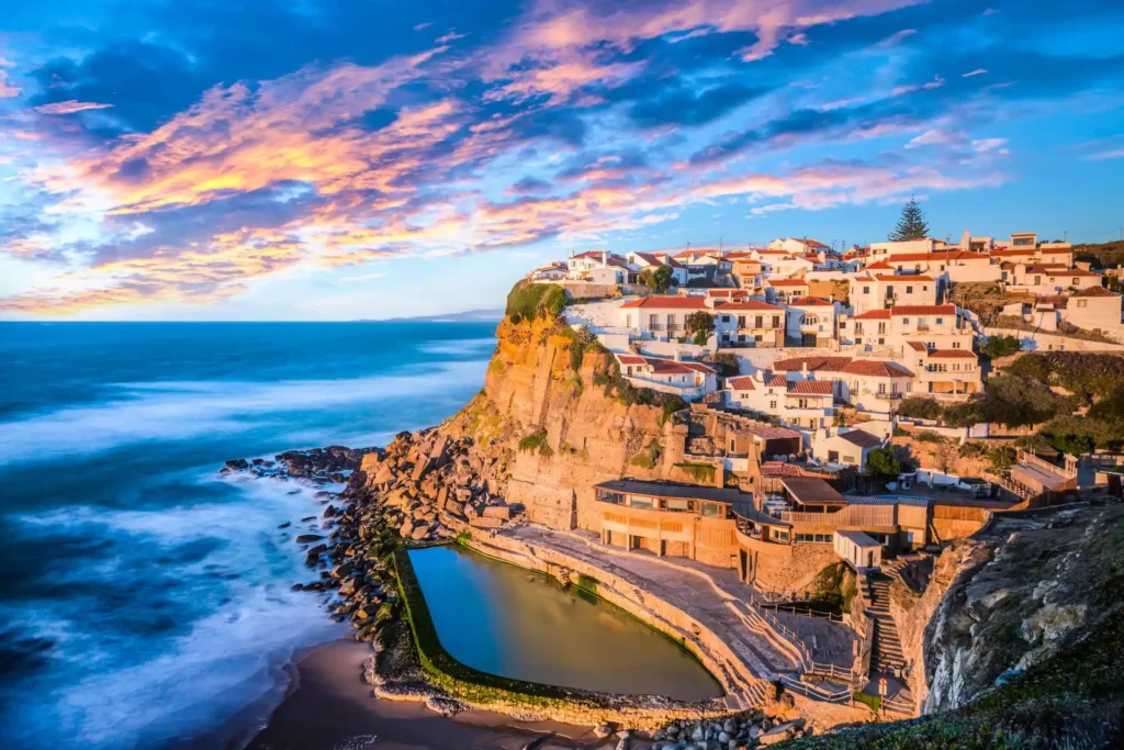 Der stimmungsvolle Küstenort Sintra bei Lissabon in Portugal bei Sonnenuntergang, mit Häusern auf einer Klippe und Blick auf das Meer und einen großen Naturpool - ein Tipp für Ihre Luxusreise nach Portugal, individuell buchbar bei der Portugal-Expertin Katharina Fenners!