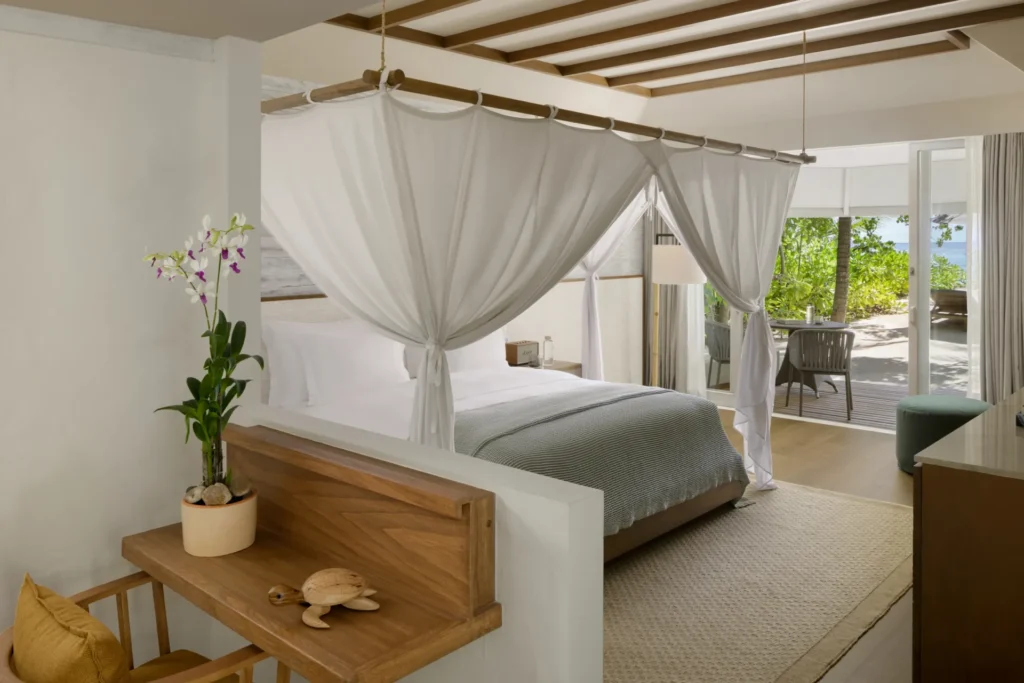 Schlafzimmer in einer Strandvilla auf den Malediven. Helle und natürliche Materialien und luftige Textilien prägen des Interior, durch die großen Fenster geht der Blick aufs Meer. Buchbar für Ihren Malediven Luxusurlaub bei Fenners Reisen!