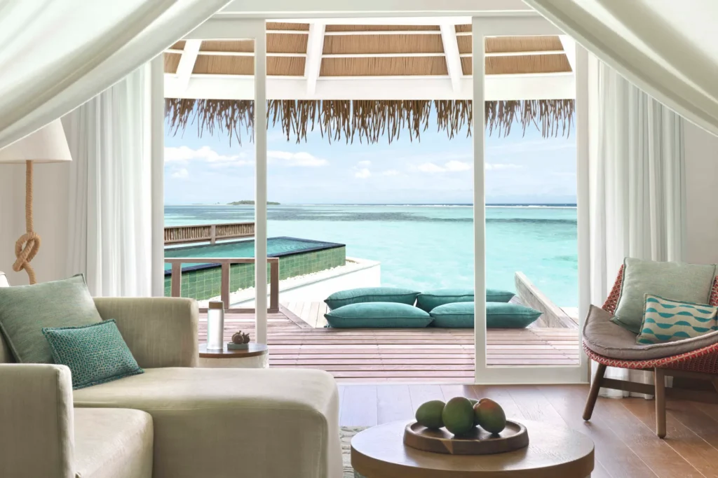Stilvoller Wohnbereich einer Wasservilla mit Pool auf den Malediven, aus den bodentiefen Fenstern geht der Blick auf die private Terrasse, den Pool und die türkise Lagune. Sie planen eine Malediven Luxusreise? Katharina Fenners kennt die besten Resorts und berät Sie individuell.