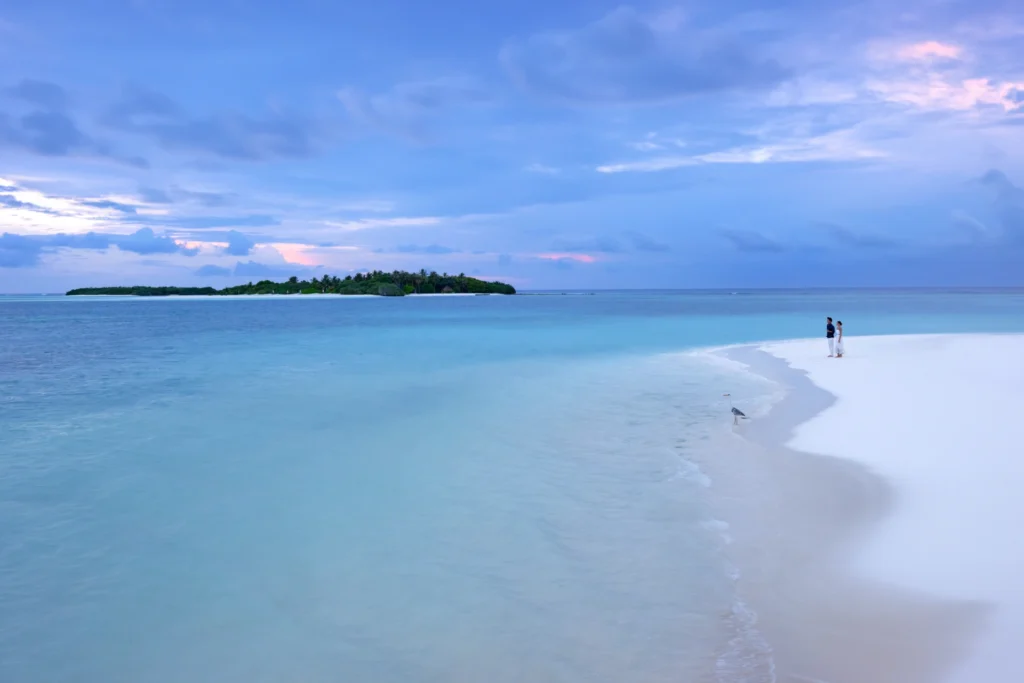 Paar im Abendlicht an einem einsamen Sandstrand mit Blick auf eine kleine Insel am Horizont - so paradiesisch ist die Kulisse nur auf den Malediven. Katharina Fenners kennt die schönsten Inseln und Resorts und berät Sie individuell!