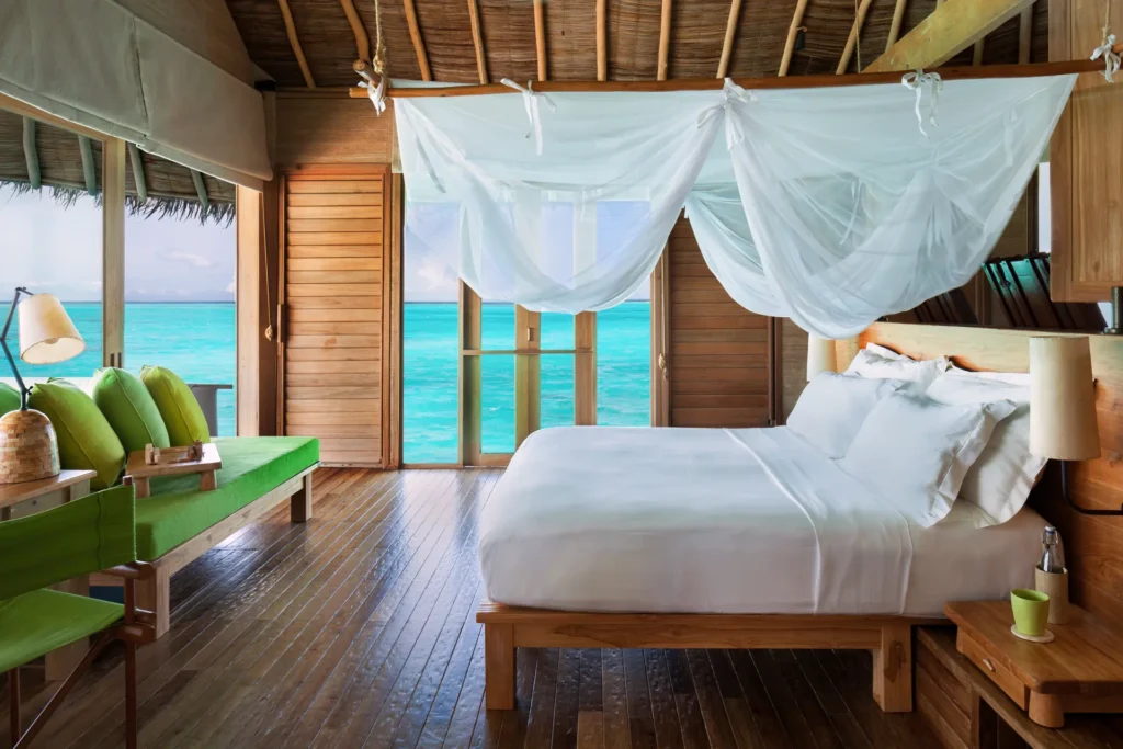 Viel Holz, weiße Textilien und ein grünes Sofa prägen das Schlafzimmer einer Laamu Wasservilla mit Pool mit weitem Rundum-Blick auf den Indischen Ozean, buchbar für eine maßgeschneiderte Maledivenreise bei Fenners Reisen!