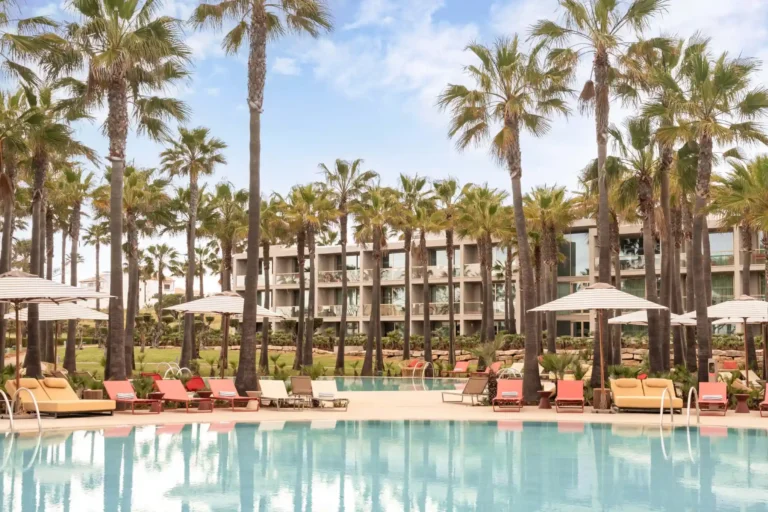 Bunte Liegestühle umrahmen den glitzernden Pool des Kimpton Atlantico Algarve Boutiquehotels, hohe Palmen umsäumen das Hotelgebäude im Hintergrund. Ein Tipp für Wellnessentuisiasten, die Wert auf Design legen - und individuell buchbar bei Fenners Reisen.