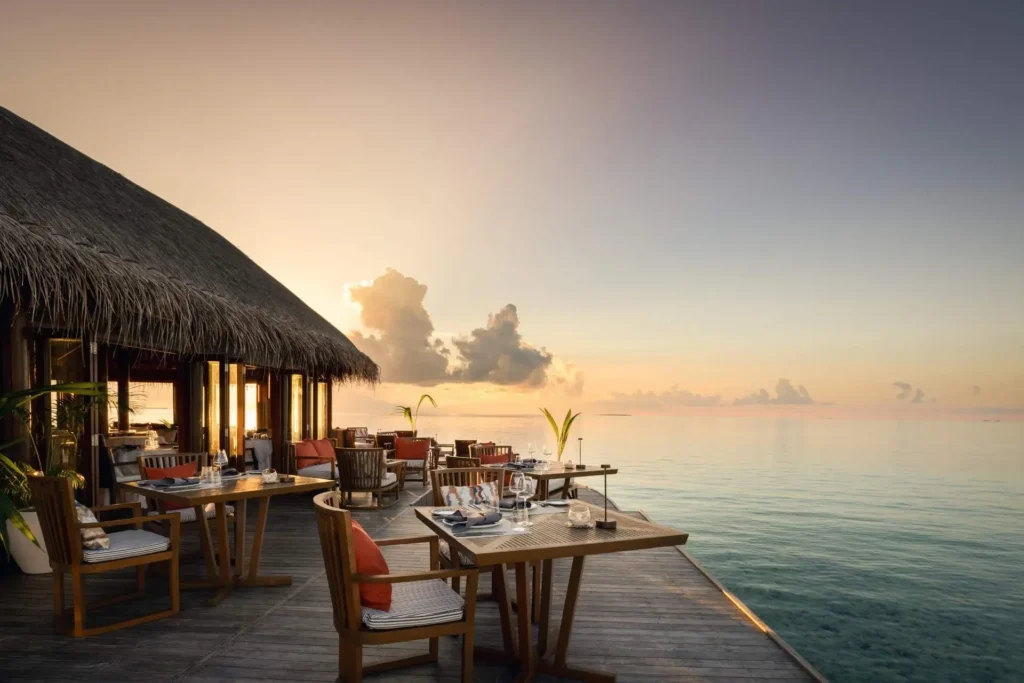 Tische auf einr Holzterrasse über dem Indischen Ozean, im Hintergrund geht die Sonne unter - Katharina Fenners kennt die besten Resorts für exklusiven Barfuß-Luxusurlaub auf den Malediven! Jetzt individuell beraten lassen.