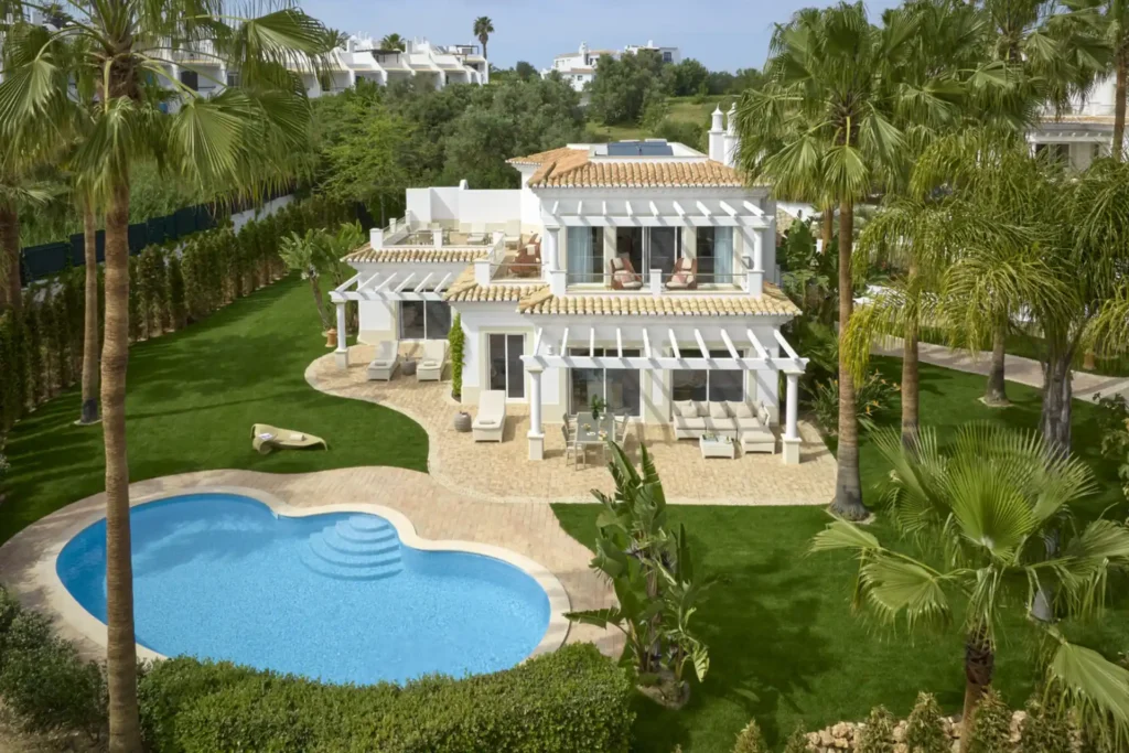 Außenansicht der zweistöckigen Vila Atlantico, umrahmt von Palmen und mit geschwungenem Lap Pool. Die Luxusvilla befindet sich in einem 5-Sterne-Resort und kann bei Fenners Reisen individuell gebucht werden - für Ihren maßgeschneiderten Luxusurlaub in Portugal.
