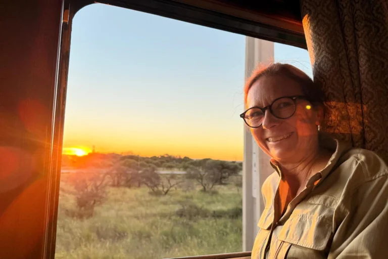 Sonnenuntergang über der Savanne Katharina Fenners lehnt am Fenster des Luxuszugs Rovos Rail, während die Sonne über der vorbeiziehenden afrikanischen Landschaft untergeht. Sie planen eine unvergessliche Reise durch den Süden Afrikas? Katharina Fenners gestaltet Ihr Abenteuer individuell!