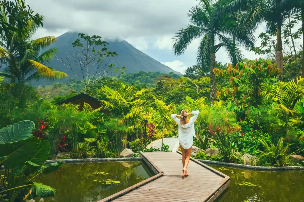 Eine Frau spaziert über einen Holzsteg zwischen den Pools des Nayara Gardens, im Hintergrund der ikonische Arenal Vulkan - buchen Sie eine unvergessliche Costa Rica Rundreise bei der Expertin Katharina Fenners!