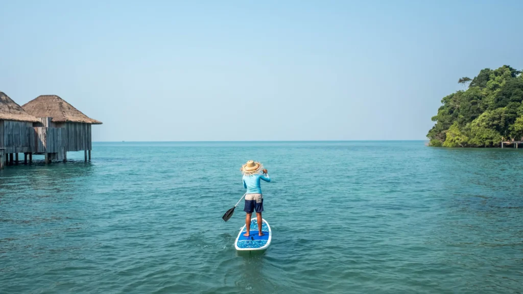 Ein Mann mit Strohhut paddelt mit dem SUP zwischen Wasservillen und einer grünen Insel über das Meer vor Kambodscha - Rundreisen durch dieses Land haben neben Kultur und Abenteuer auch traumhafte Bilderbuchstrände zu bieten! Katharina Fenners gestaltet Ihre Reise durch Kambodscha individuell.